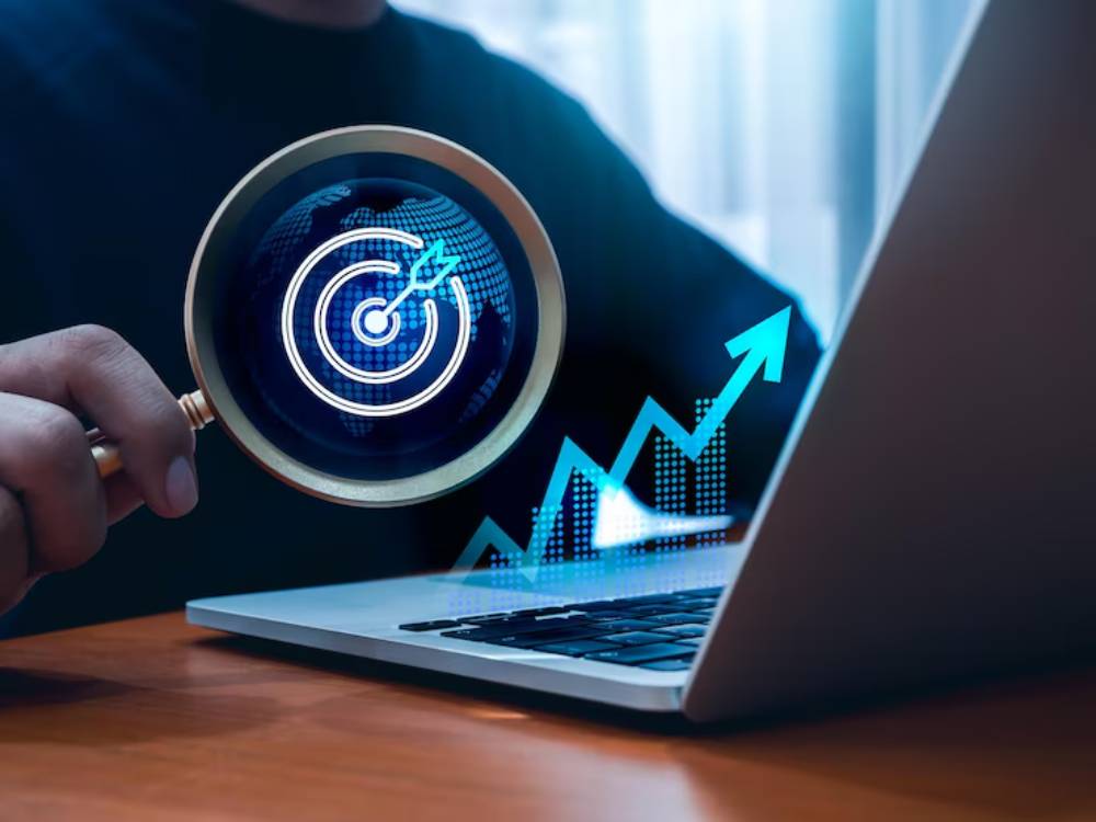 A person holding a magnifying glass over a laptop, with a digital target and upward graph symbolizing data analysis and goal setting.