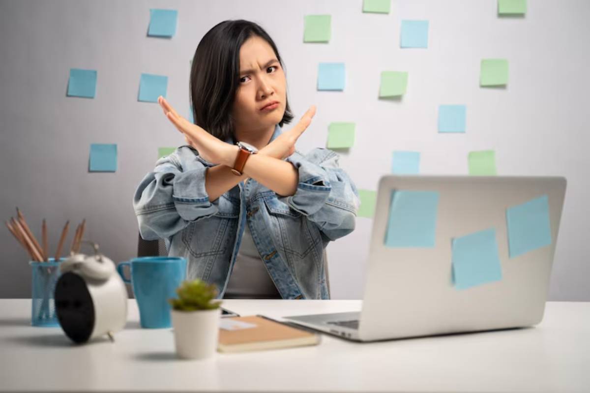 A person in a denim jacket crosses their arms in front of a desk with a laptop, stationary, and a plant, surrounded by colorful sticky notes. 