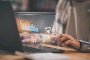 Person typing on a laptop with a transparent overlay showing graphs and a shopping cart icon. The scene suggests e-commerce and data analysis.