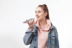 A young woman in a denim jacket and pink hoodie holds a smartphone to her ear, appearing giving voice command.