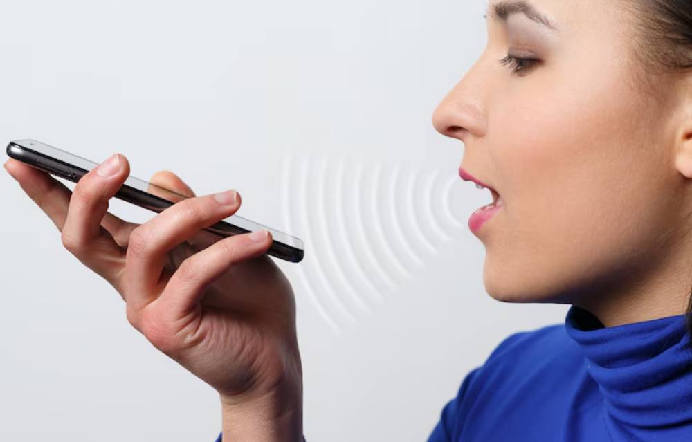 A person holds a smartphone to their mouth, speaking into it, with sound waves radiating from the device against a light background.