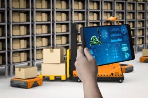 A hand holds a tablet displaying warehouse data, while automated vehicles and stacked boxes fill the background.