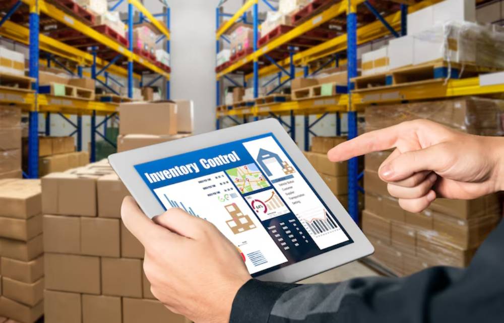 A person in a warehouse uses a tablet displaying inventory control data while surrounded by stacked boxes and shelves.