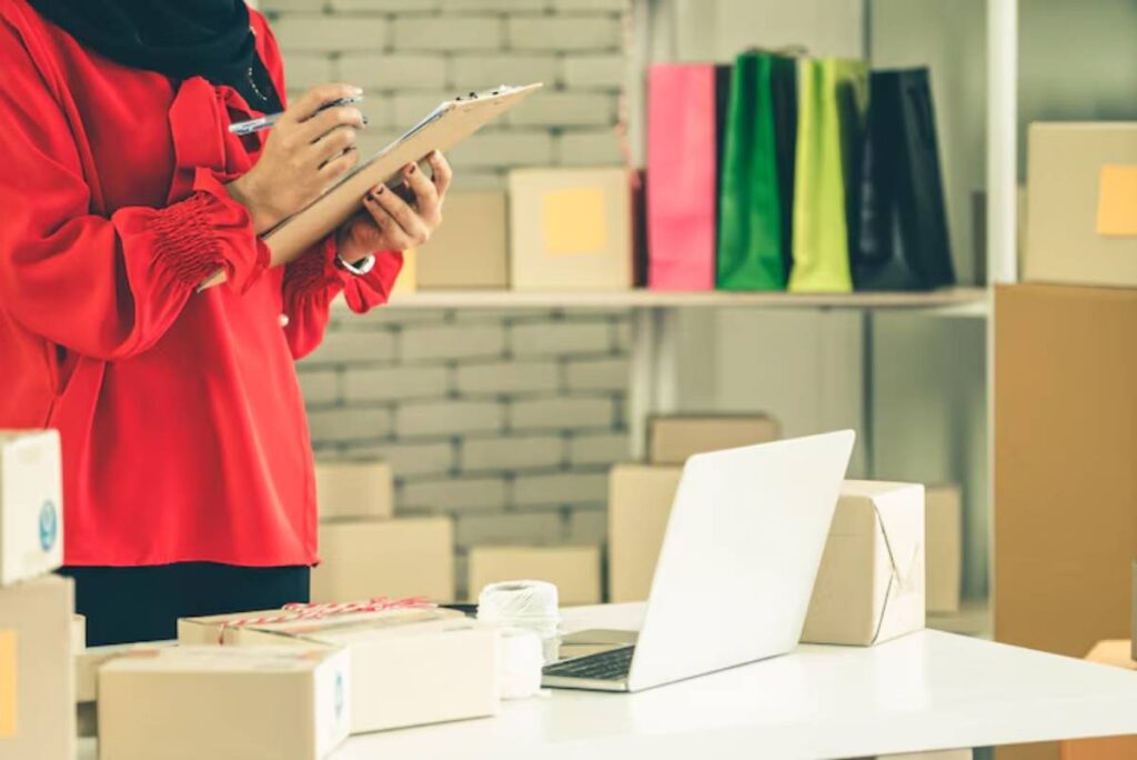 A person in a red shirt holds a clipboard with a pen, surrounded by boxes and a laptop in a workspace filled with colorful bags.