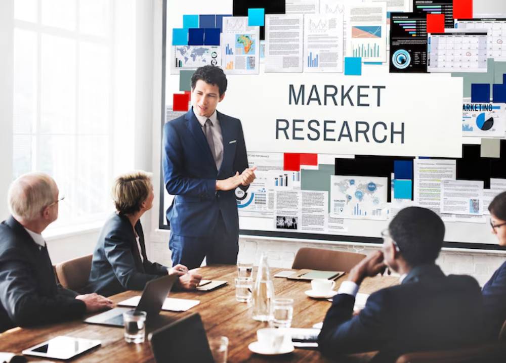 A professional meeting room with a presenter discussing market research, surrounded by engaged attendees and charts on display.