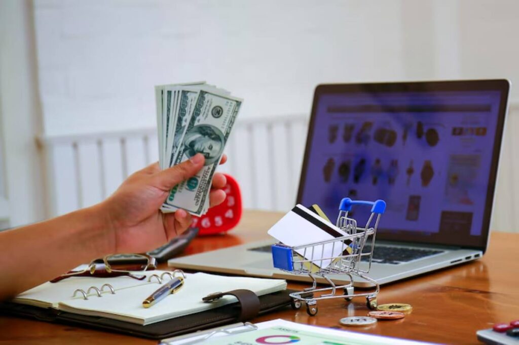 A hand holding cash next to a laptop displaying shopping website, with a mini shopping cart and notebook on a wooden desk.