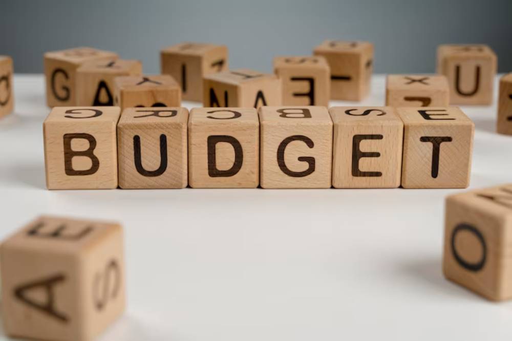 Wooden blocks arranged to spell 'BUDGET,' surrounded by scattered alphabet blocks on a white surface.