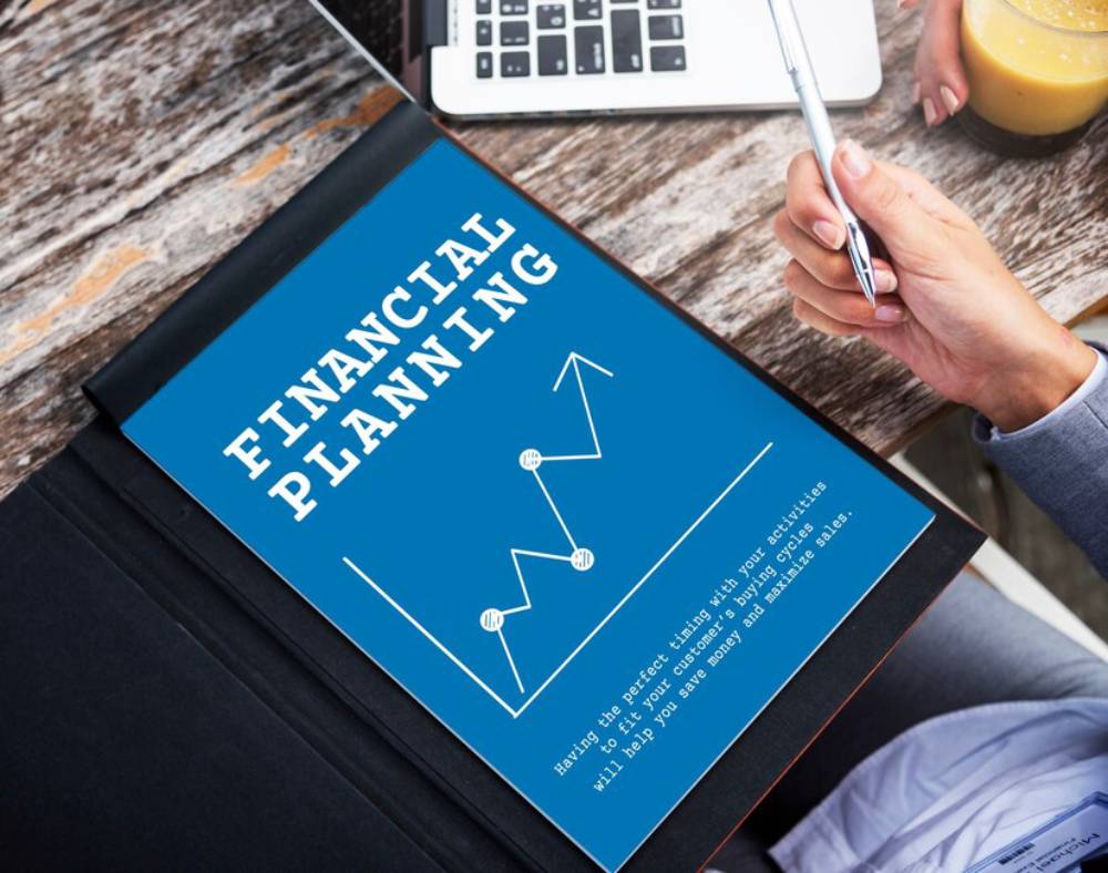 A person holds a pen over a financial planning document featuring a graph, with a laptop and drink nearby on a wooden table.