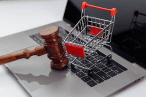 A wooden gavel rests on a laptop keyboard beside a small metal shopping cart, symbolizing online commerce and legal themes.