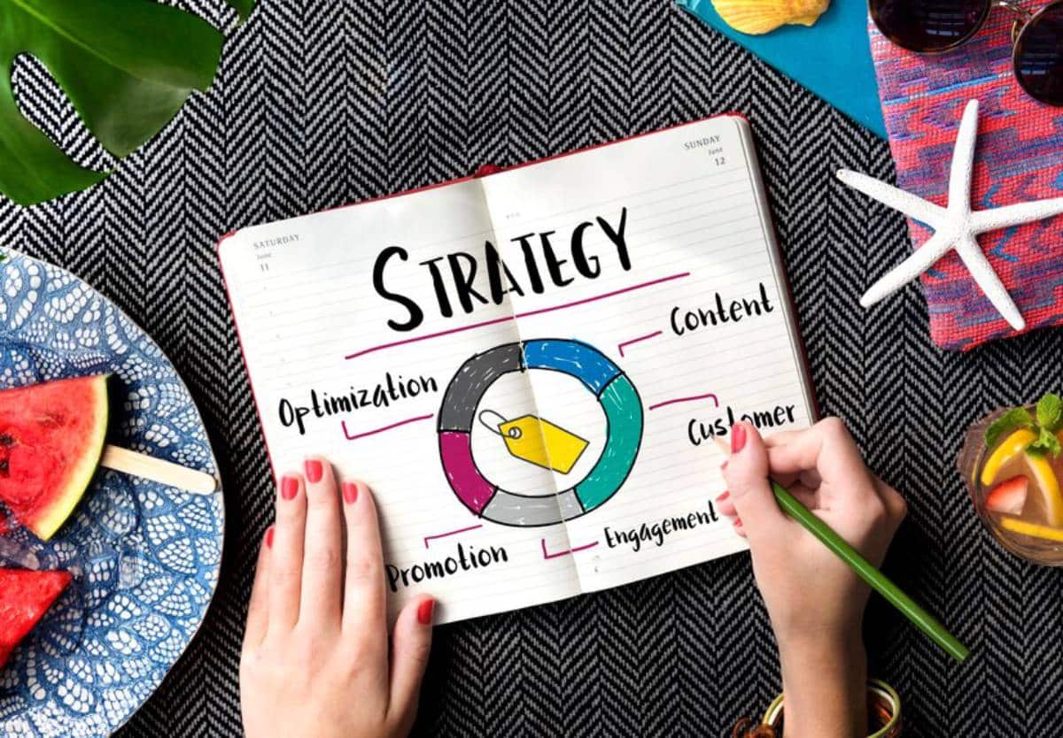 A hand holding a green pen writes about a strategy diagram in a notebook, surrounded by tropical elements like watermelon and drinks.