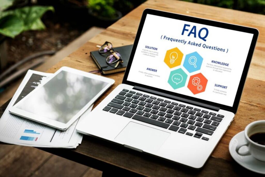 A laptop displaying a FAQ infographic, surrounded by a tablet, glasses, documents, and a coffee cup on a wooden table.