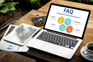 A laptop displaying a FAQ infographic, surrounded by a tablet, glasses, documents, and a coffee cup on a wooden table.