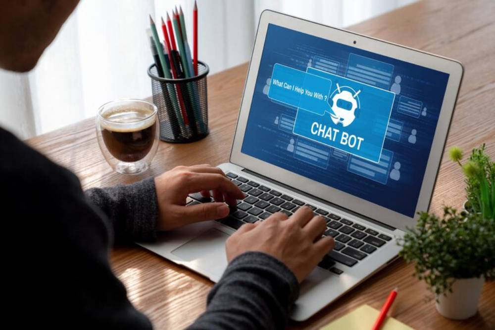 A person types on a laptop displaying a chatbot interface, with coffee and pencils nearby, in a bright workspace.
