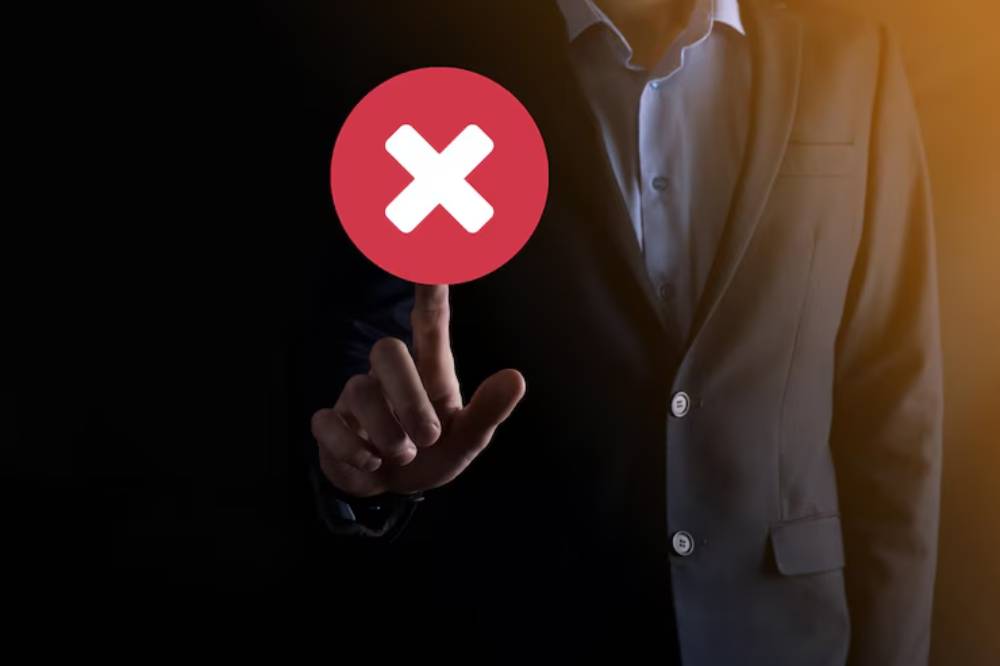 A person in a suit, partially visible, reaches towards a floating red circle with a white 'X,' symbolizing error or denial, set against a dark background. 