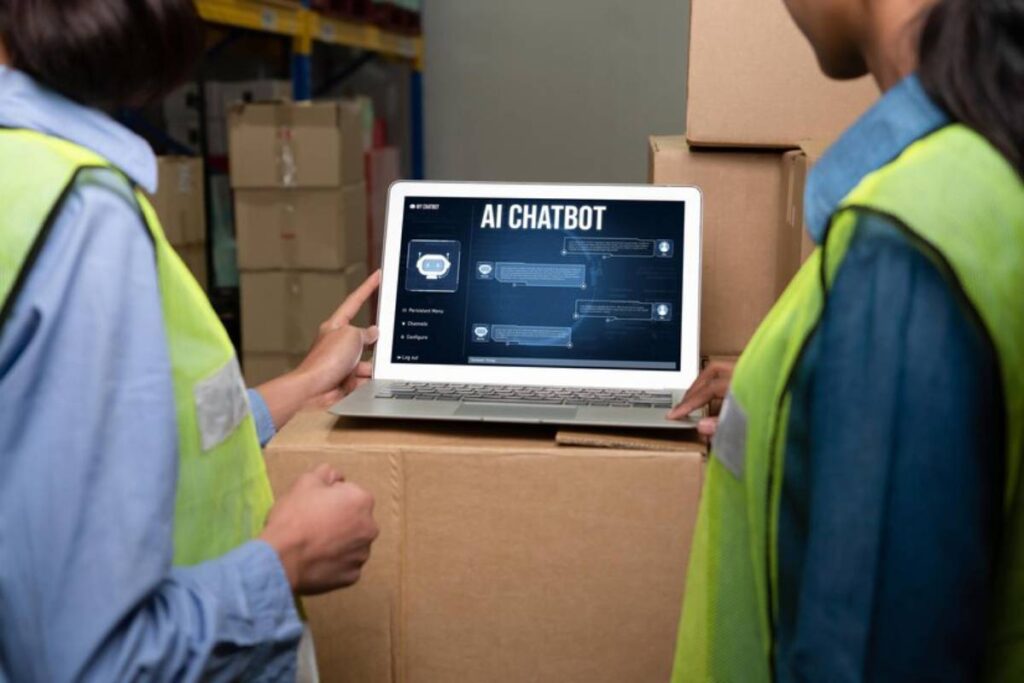 Two warehouse workers in safety vests review an AI chatbot interface on a laptop positioned on stacked cardboard boxes.
