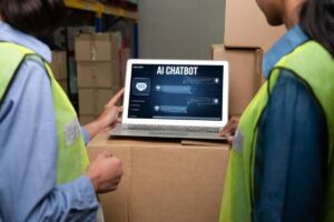Two warehouse workers in safety vests review an AI chatbot interface on a laptop positioned on stacked cardboard boxes.