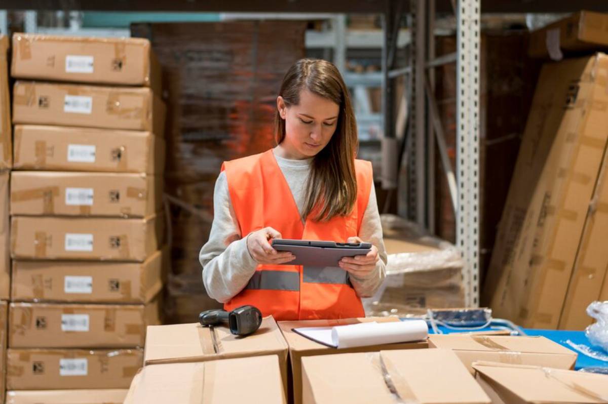 A woman in an orange safety vest stands in a warehouse, using a tablet while surrounded by stacked cardboard boxes.