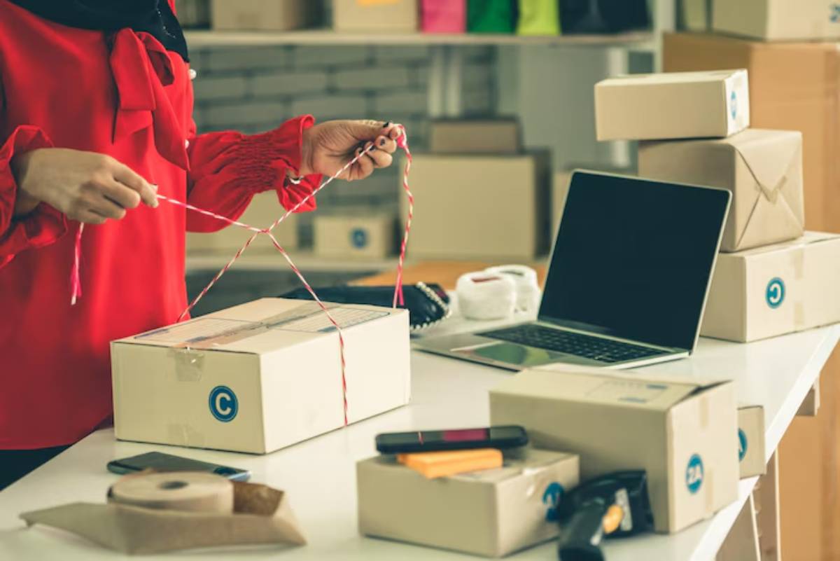 A woman in a red blouse ties a string around a package at a desk with various shipping supplies and a laptop nearby.