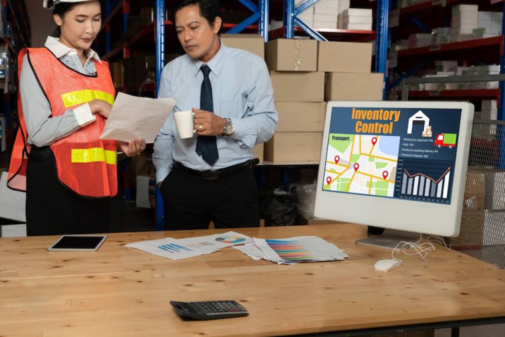 Two professionals discuss documents in a warehouse, with a computer displaying inventory control graphics and charts on a wooden table.