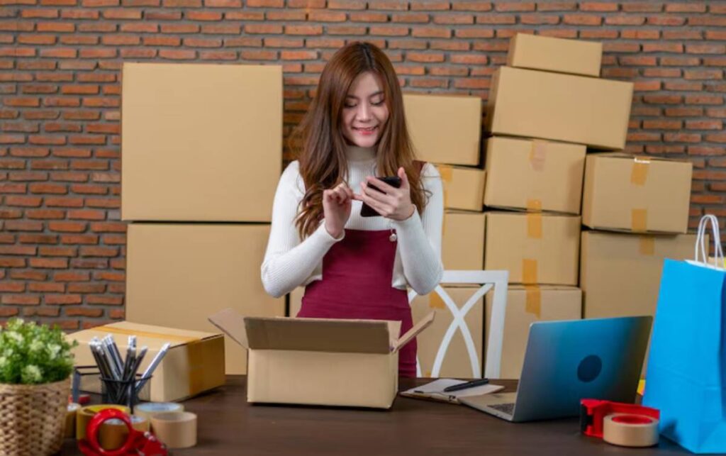 A woman in a white sweater and burgundy dress checks her phone while standing by a table with an open box and packing materials.