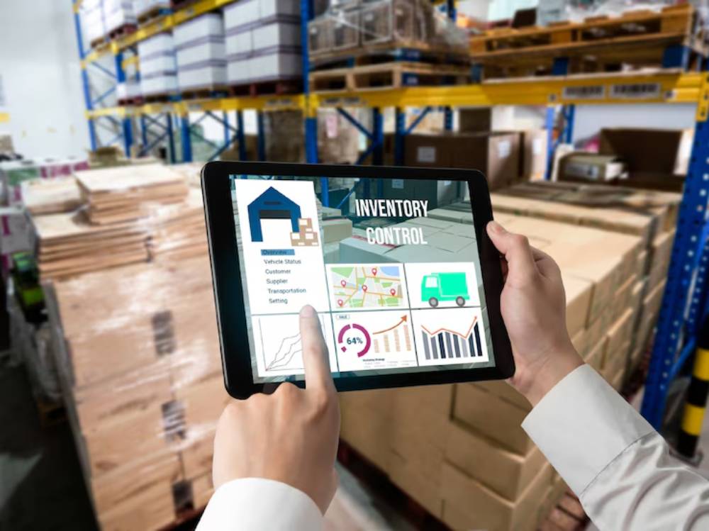 A person holds a tablet displaying an inventory control interface, with stacks of boxes in a warehouse background.