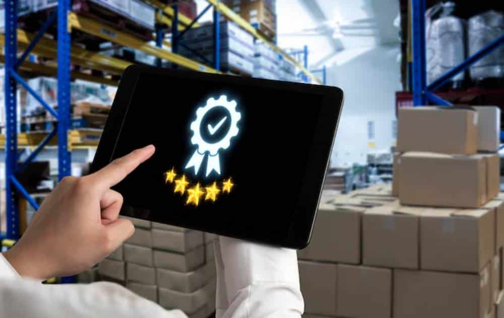 A hand points at a tablet displaying a glowing badge with a checkmark and five stars, set against a warehouse background with stacked boxes.