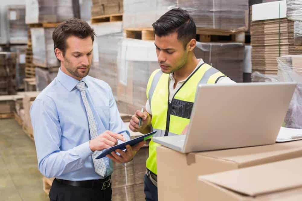 Two men discuss inventory in a warehouse, with stacks of pallets and cardboard boxes in the background; a laptop is open on the table.