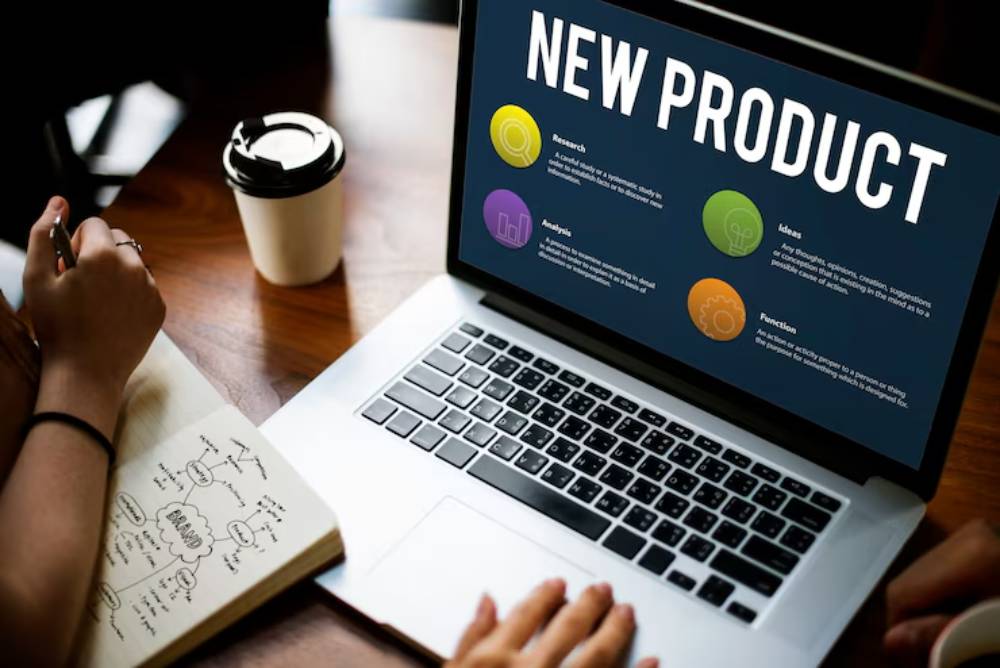 A laptop displaying 'New Product' with sections for Research, Analysis, Ideas, and Function, beside a notebook and coffee cup.