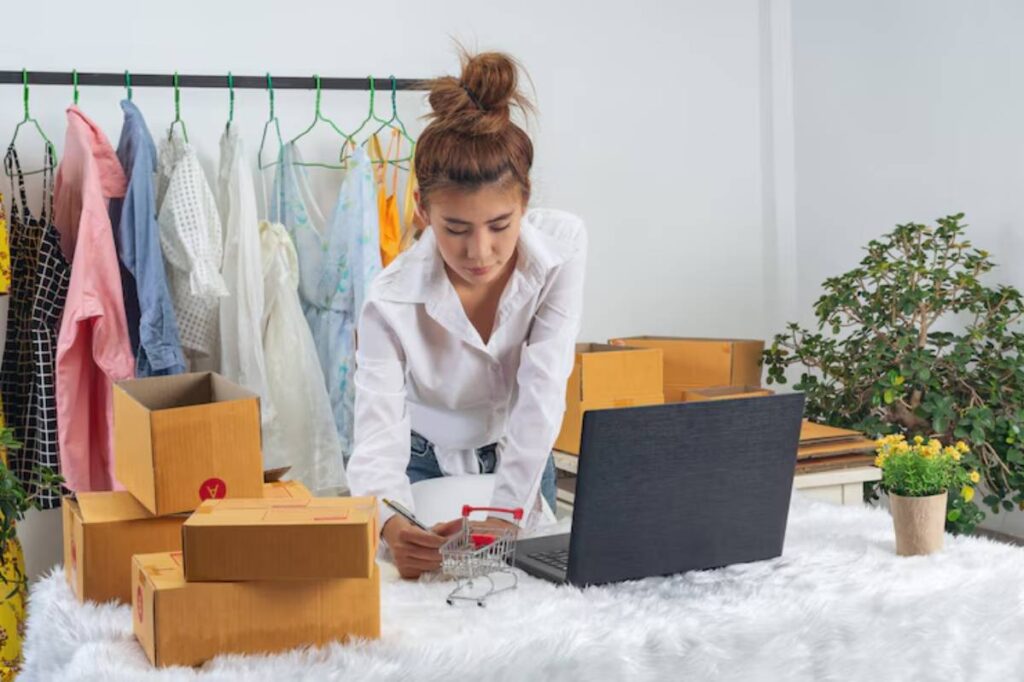 A person in a white shirt stands over a laptop, surrounded by cardboard boxes and colorful clothing on a rack, with a small plant nearby.