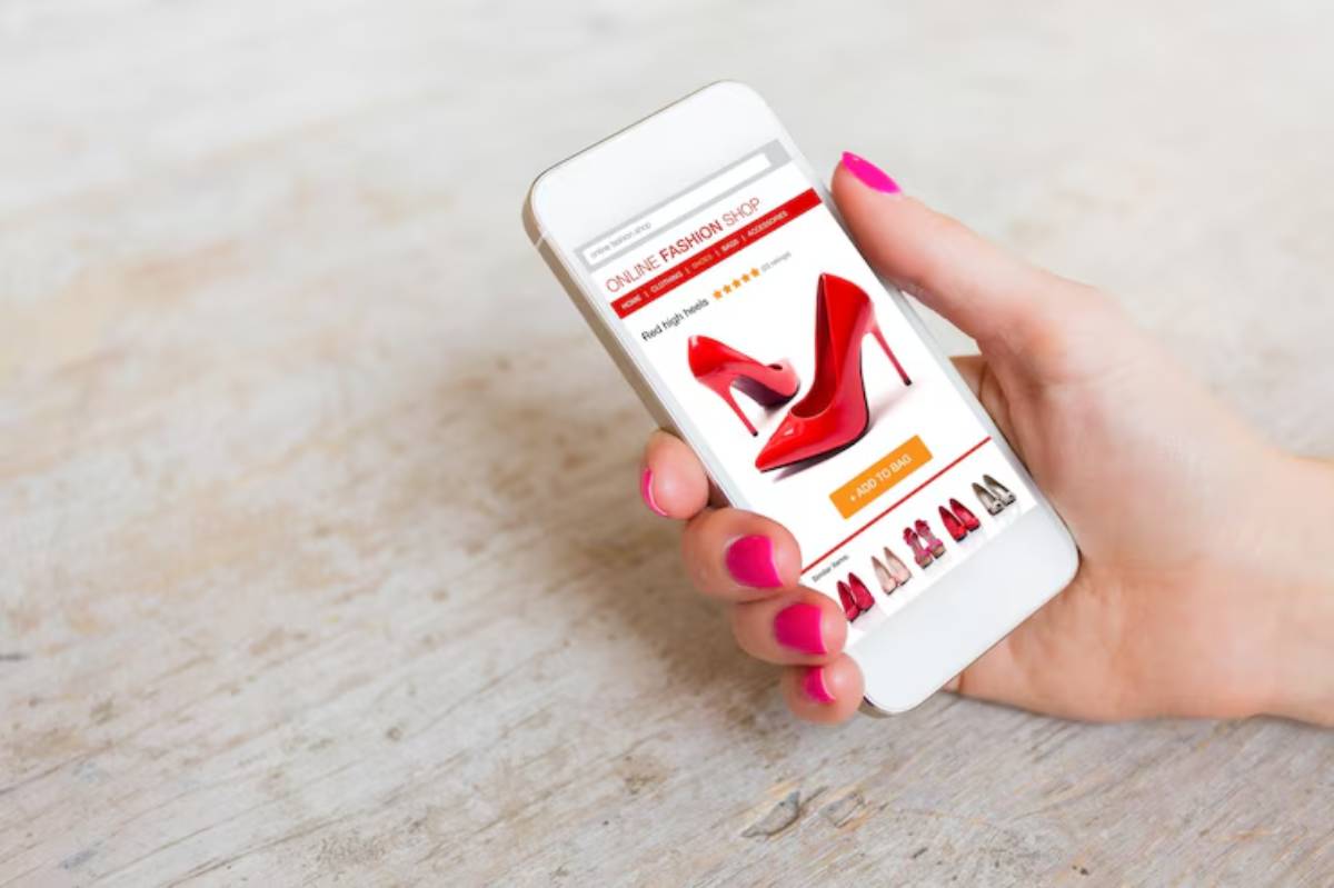 A hand with pink nails holds a smartphone displaying a red high heel on a fashion shopping app.