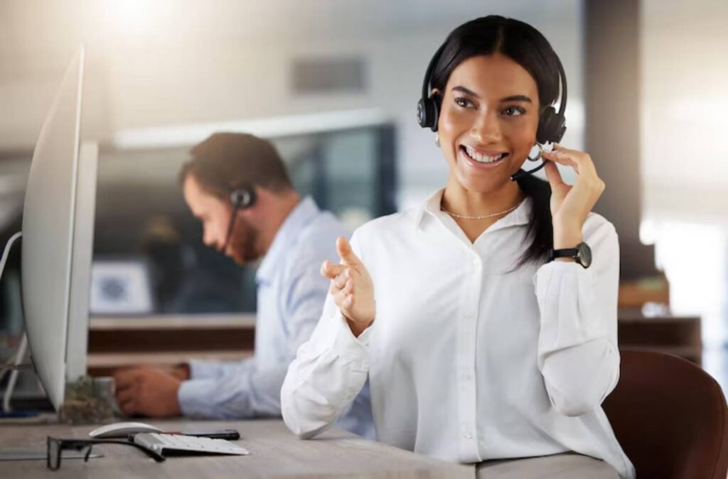 A woman wearing headphones gestures while speaking on a headset at a modern office desk, with a blurred coworker in the background.