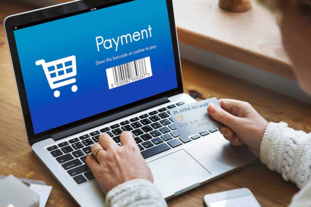 A person holding a credit card above a laptop displaying a payment screen with a shopping cart and barcode for checkout.