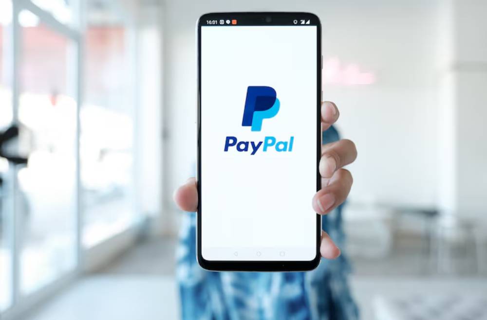 A person holds a smartphone displaying the PayPal logo against a bright, modern interior background.