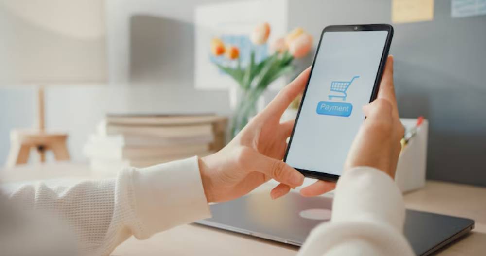 A person using a smartphone to make an online payment, displaying a shopping cart icon and the word 'Payment' on the screen.
