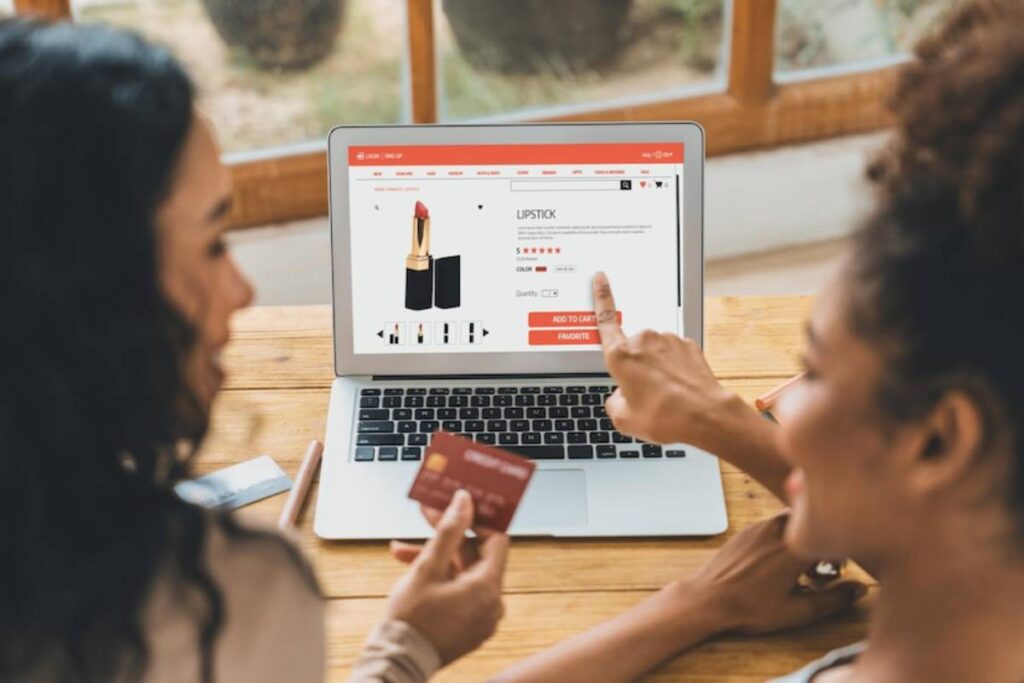 Two people are shopping online for lipstick, pointing at a laptop screen displaying product details while holding credit cards.