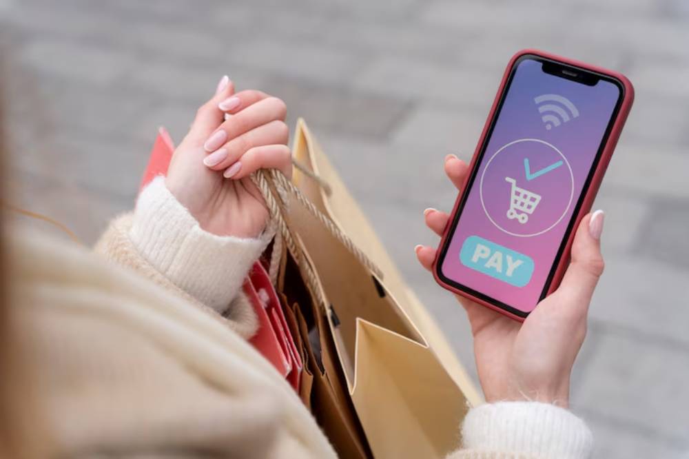 A hand holding a smartphone displaying a 'PAY' button and shopping cart icon, set against shopping bags on a street.