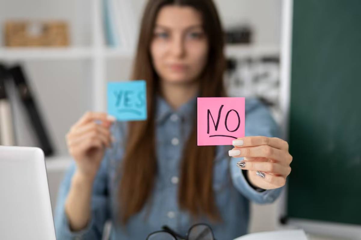 A person holds two sticky notes, one blue with 'YES' and one pink with 'NO,' sitting at a desk in an office setting.