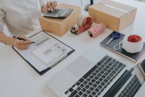 A person calculates shipping costs at a desk with boxes, packing tape, scissors, labels, and a laptop.
