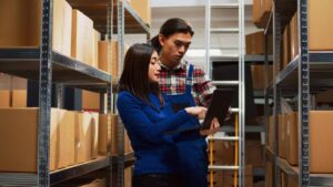 Two individuals collaborate in a warehouse, reviewing information on a laptop amidst shelves of neatly stacked cardboard boxes.