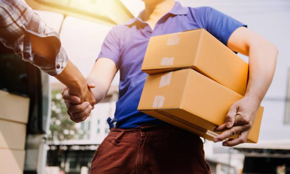 A delivery person in a blue shirt shakes hands with a customer while holding two brown boxes in the other hand.