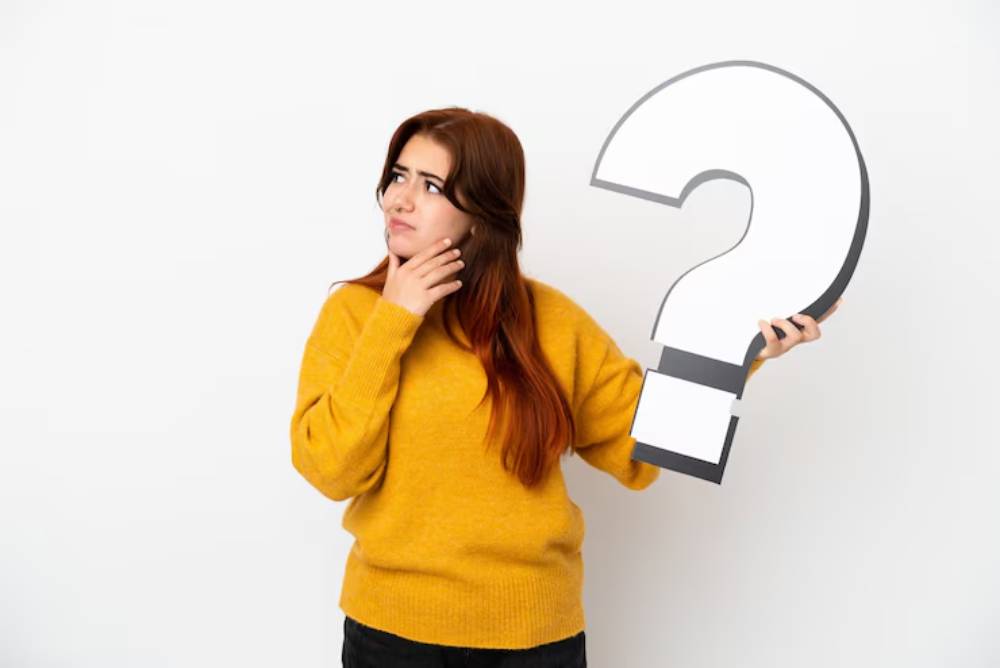 A woman wearing a yellow sweater holds a large question mark in front of a white background, appearing thoughtful and curious.