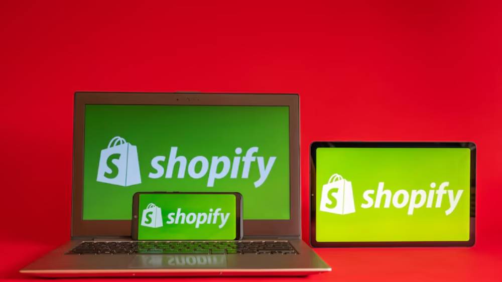 Three devices (laptop, tablet, smartphone) display the Shopify logo on vibrant green screens, set against a red background.