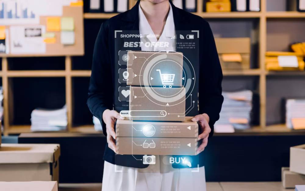 A businesswoman holds stacked boxes while holographic shopping icons and offers appear in front of her in a modern workspace.